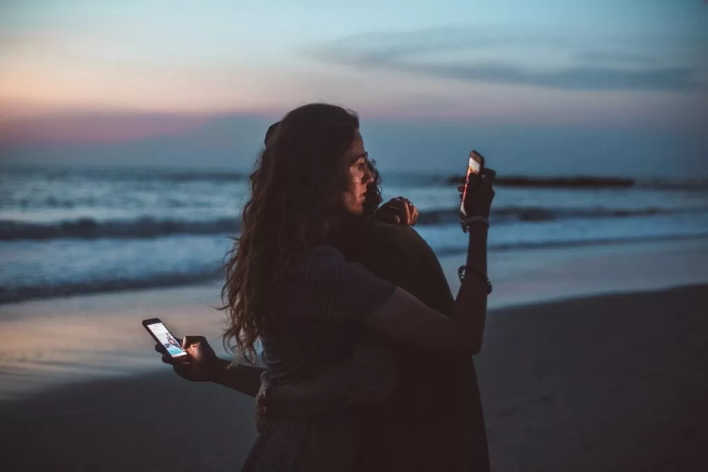 Two people hugging each other on the beach but looking on their phones which brings a disconnection feeling